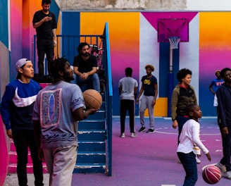 A vibrant outdoor basketball court filled with people of various ages engaged in different activities. Some are playing basketball while others are standing and chatting. The court is surrounded by brightly painted walls in shades of pink, orange, and blue. A basketball hoop is mounted on the back wall, adding to the sports theme. A few people are sitting on stairs or using phones, creating a relaxed and social atmosphere.