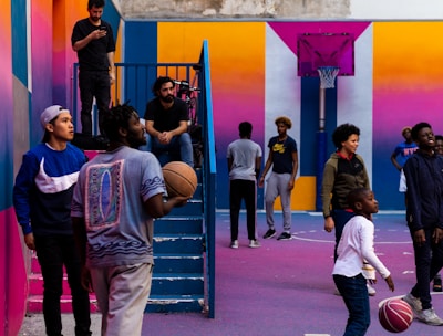 A vibrant outdoor basketball court filled with people of various ages engaged in different activities. Some are playing basketball while others are standing and chatting. The court is surrounded by brightly painted walls in shades of pink, orange, and blue. A basketball hoop is mounted on the back wall, adding to the sports theme. A few people are sitting on stairs or using phones, creating a relaxed and social atmosphere.