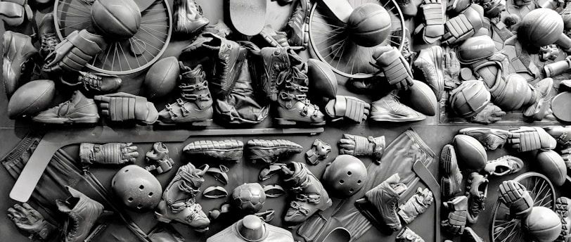 A complex arrangement of various sports equipment and attire, including tennis rackets, soccer balls, basketballs, helmets, shoes, gloves, and a mannequin torso wearing a T-shirt. The items are layered and artistically displayed in a monochrome color scheme, creating an intricate texture and visual pattern.