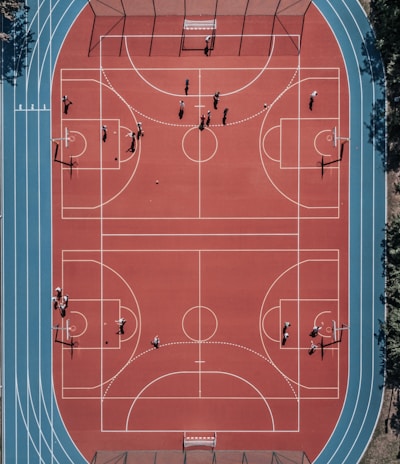 An aerial view of a large, vibrant sports complex featuring a red basketball court with multiple hoops and a surrounding blue running track. Several people are scattered across the area, engaged in various activities.