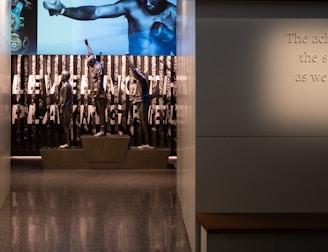 A dimly lit display features statues of athletes standing on a podium. They are positioned in front of a backdrop with the words 'LEVELING THE PLAYING FIELD' in bold letters. On the wall to the right, a softly lit quote is partially visible. The overall ambiance suggests a museum or exhibition setting.