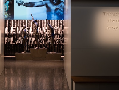 A dimly lit display features statues of athletes standing on a podium. They are positioned in front of a backdrop with the words 'LEVELING THE PLAYING FIELD' in bold letters. On the wall to the right, a softly lit quote is partially visible. The overall ambiance suggests a museum or exhibition setting.