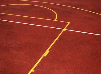 A sports court with a reddish-brown surface and bright yellow and white lines. The lines form geometric shapes, including rectangles and circles, suggesting markings for games like basketball or netball.