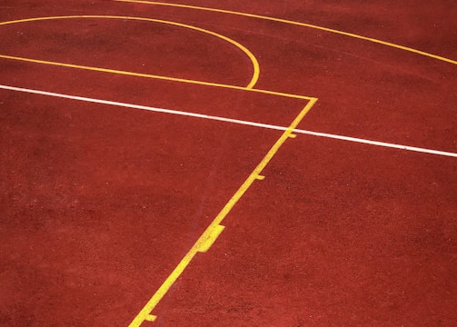 A sports court with a reddish-brown surface and bright yellow and white lines. The lines form geometric shapes, including rectangles and circles, suggesting markings for games like basketball or netball.