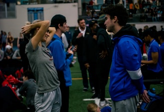 People engage in conversation and activities in an indoor sports facility. A woman in gray athletic wear stretches her arms above her head while talking to a man in a blue jacket. Other people are seen interacting in the background, some are seated on the ground, and the atmosphere is lively with many individuals gathered.