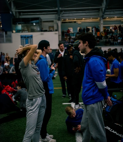 People engage in conversation and activities in an indoor sports facility. A woman in gray athletic wear stretches her arms above her head while talking to a man in a blue jacket. Other people are seen interacting in the background, some are seated on the ground, and the atmosphere is lively with many individuals gathered.