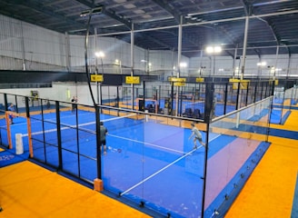 An indoor sports facility featuring several padel courts with blue playing surfaces and orange sidelines. There are players engaged in a game on one of the courts, while the others appear empty. The area is well-lit with overhead lighting and enclosed by a high ceiling and metal structure.