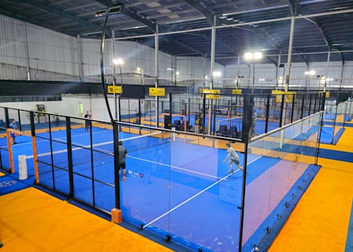 An indoor sports facility featuring several padel courts with blue playing surfaces and orange sidelines. There are players engaged in a game on one of the courts, while the others appear empty. The area is well-lit with overhead lighting and enclosed by a high ceiling and metal structure.