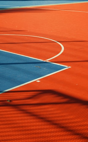 A vibrant sports court with sections of red and blue, featuring crisp white lines marking the boundaries. Shadows from a structure create geometric patterns across the surface, adding depth and interest to the scene.